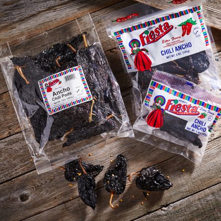 Packages of Fiesta Brand Ancho Chili Pods on a wooden surface, alongside a few loose dried chilies. Perfect for adding rich, smoky flavors to chili recipes.