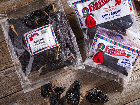 Packages of Fiesta Brand Ancho Chili Pods on a wooden surface, alongside a few loose dried chilies. Perfect for adding rich, smoky flavors to chili recipes.